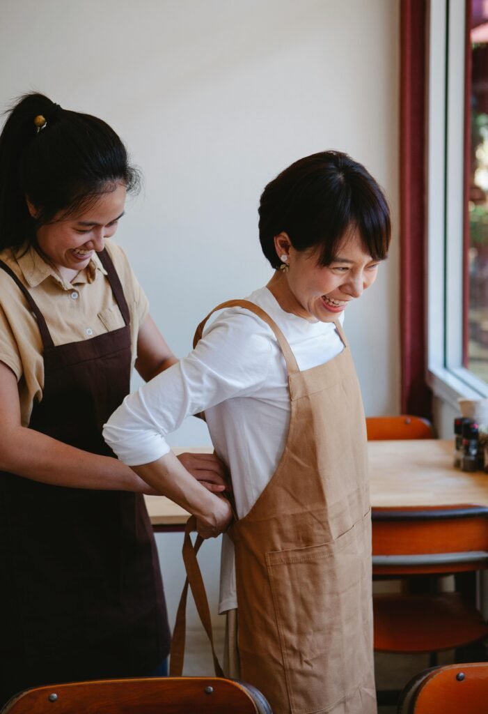 Two Asian women in aprons smiling as they help each other in a restaurant setting.
